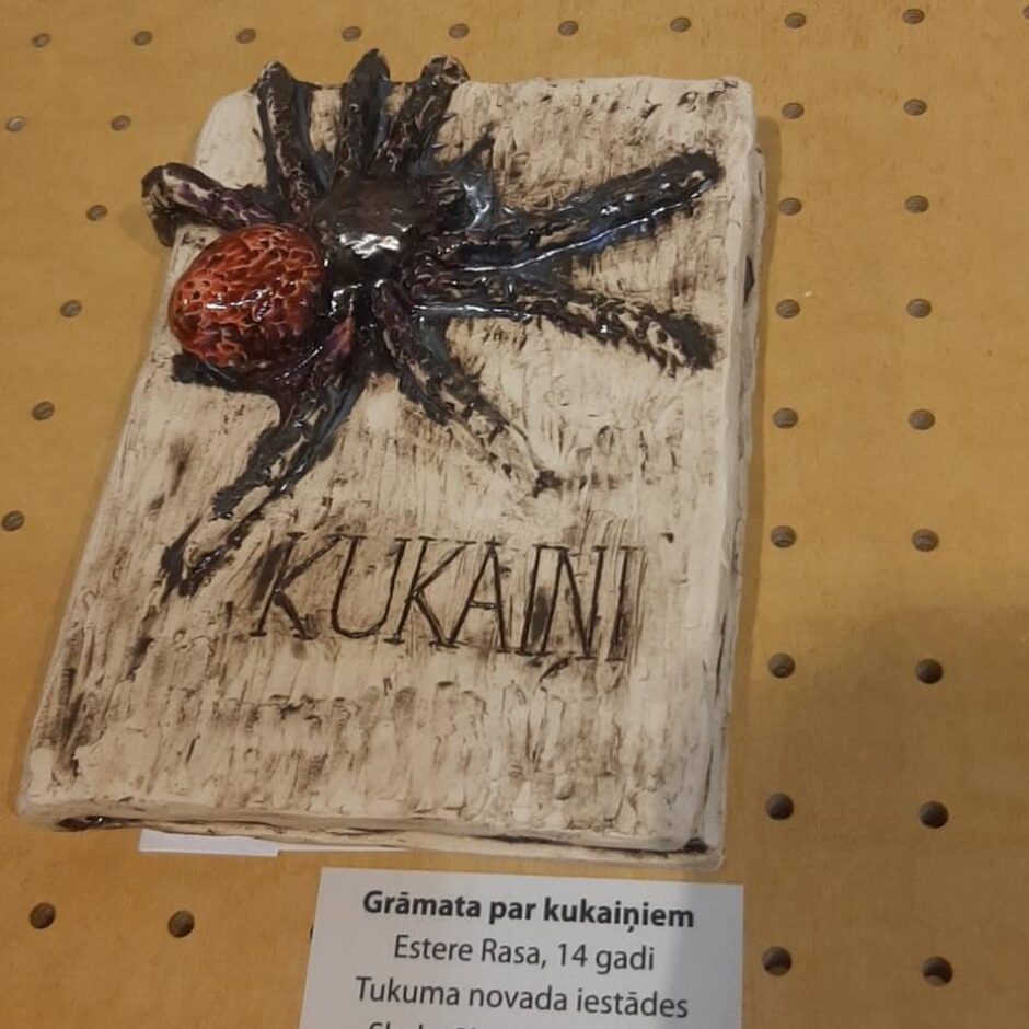 Gaišs keramikas darbs grāmatas formā ar tumšu, sarkanīgu zirnekli uz vāka un iegravētu vārdu 