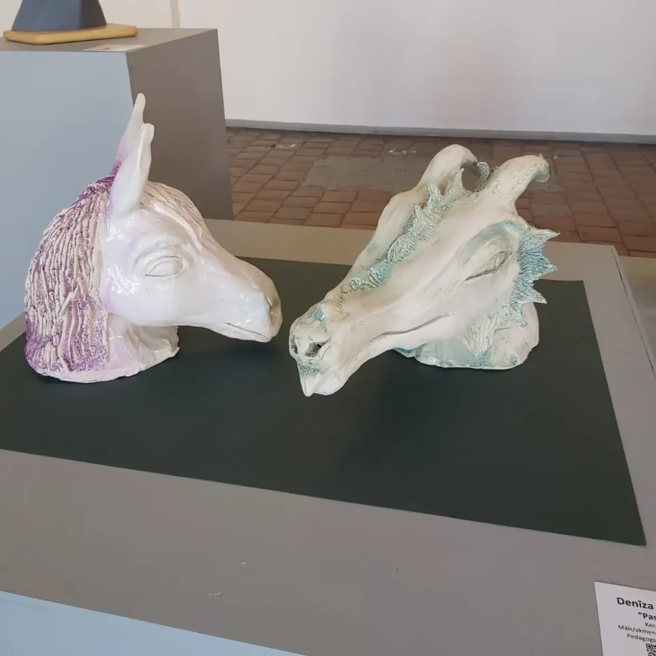 Divas keramikas skulptūras: balta zirgam līdzīga galva ar violetām krēpēm un balta pūķim līdzīga galva ar gaiši zilām detaļām, vērstas viena pret otru.